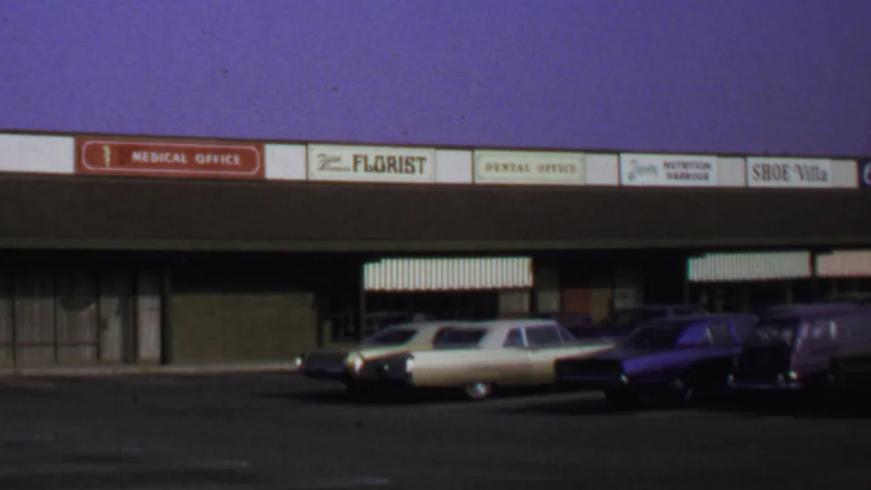 Wide home-movie frame of a Washington, DC commercial strip with multiple signs, striped awnings, and late-1960s cars parked outside.