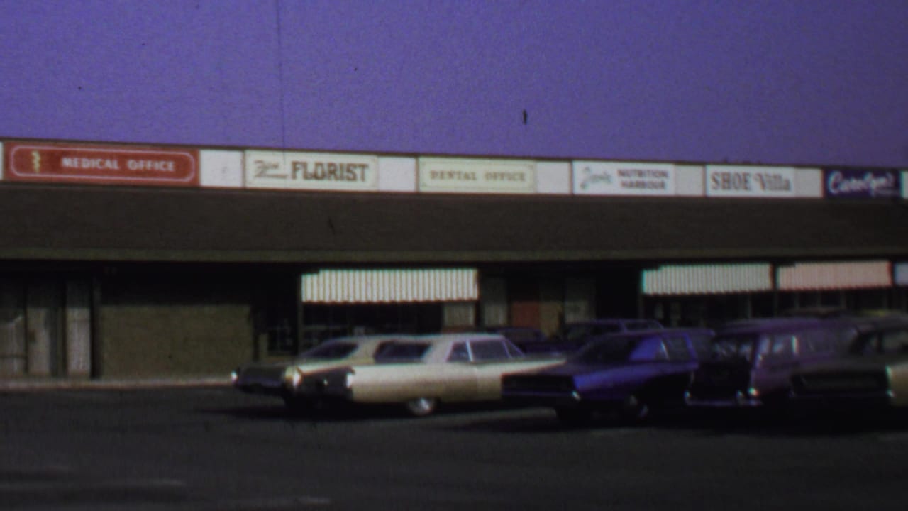 Washington DC 1969 home-movie frame of parked classic cars facing a retail strip with period shop signs.