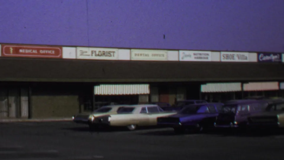 Washington DC 1969 home-movie frame of parked classic cars facing a retail strip with period shop signs.
