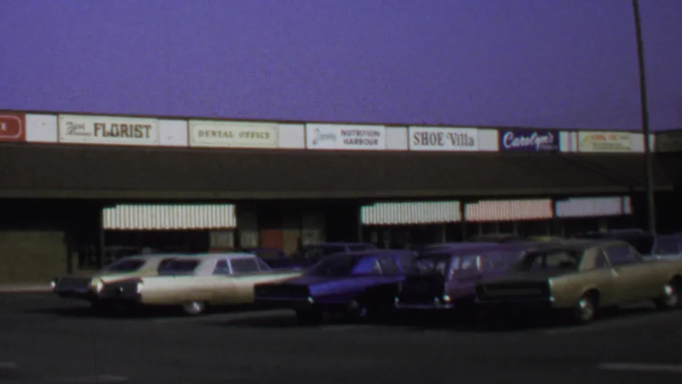 Late-1960s Washington DC 8mm frame with storefront signage, awnings, and parked American cars in a shopping-center lot.