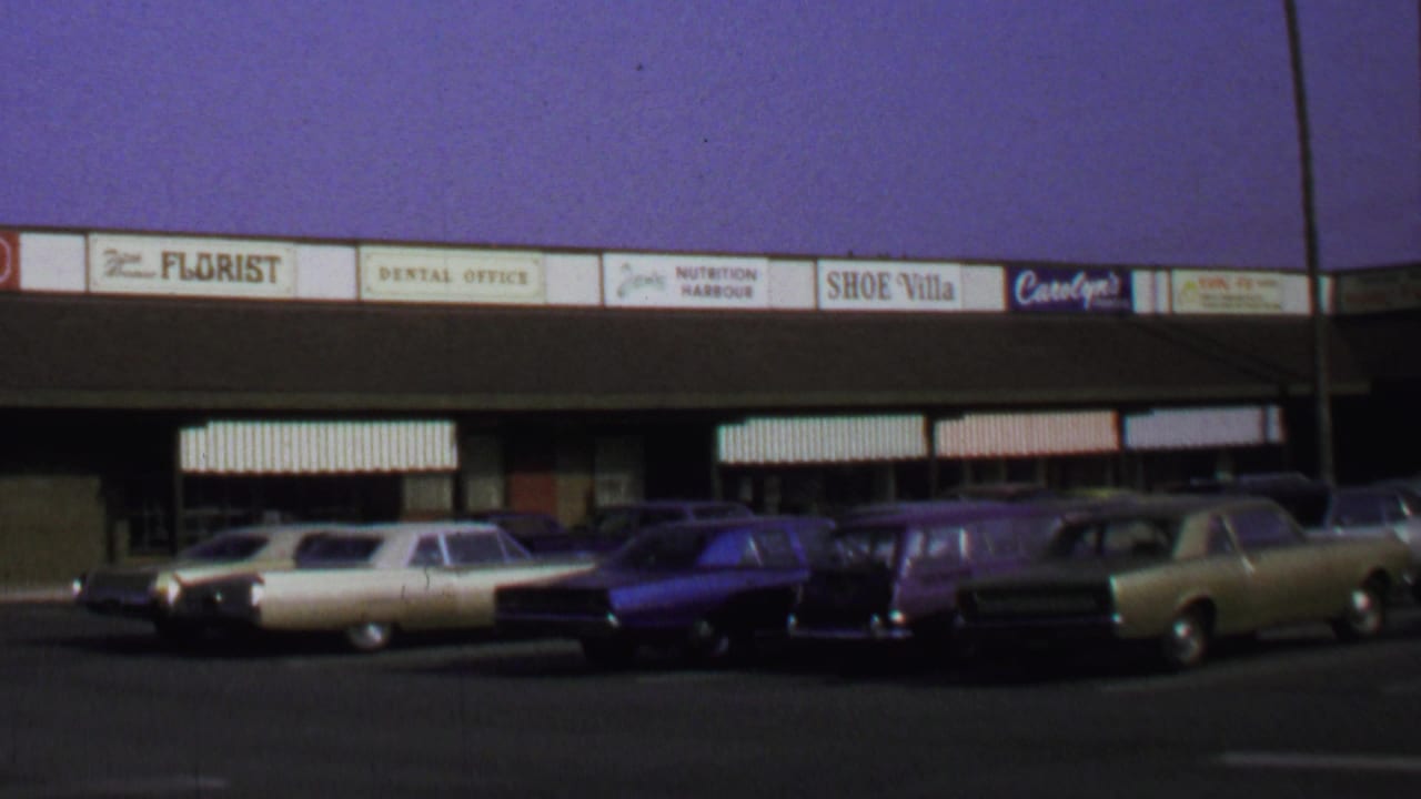 1969 Washington DC 8mm frame showing parked sedans and wagons across from storefronts including a florist, dental office, and shoe shop.