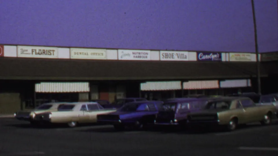 1969 Washington DC 8mm frame showing parked sedans and wagons across from storefronts including a florist, dental office, and shoe shop.