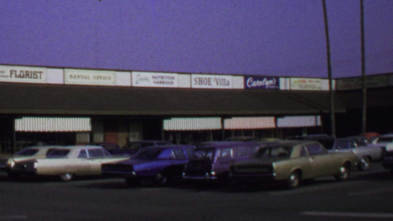 1969 Washington DC frame with cream, dark, and gold cars parked before a commercial strip with readable shop signs and striped awnings.