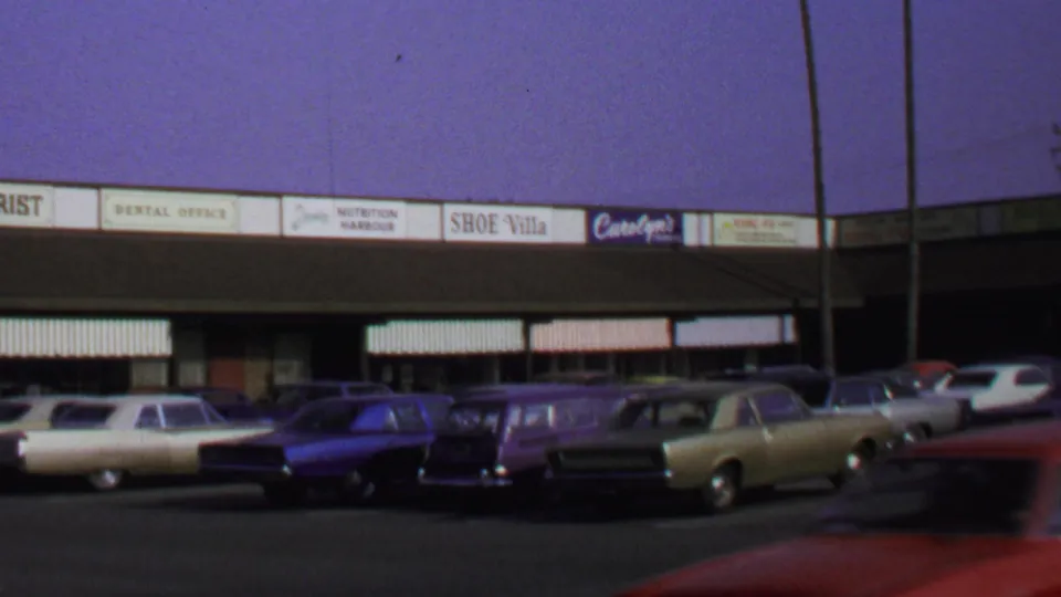 1969 Washington DC 8mm frame showing parked cars at storefronts while a red vehicle passes blurred across the parking lot foreground.