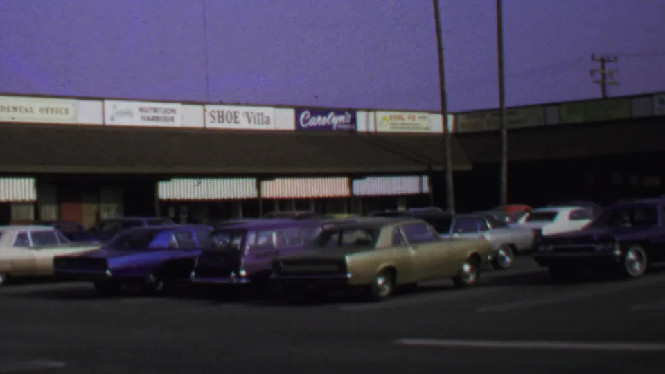 1969 Washington DC frame showing a wider parking-lot view of vintage cars, storefront signage, awnings, and utility poles at a low shopping strip.