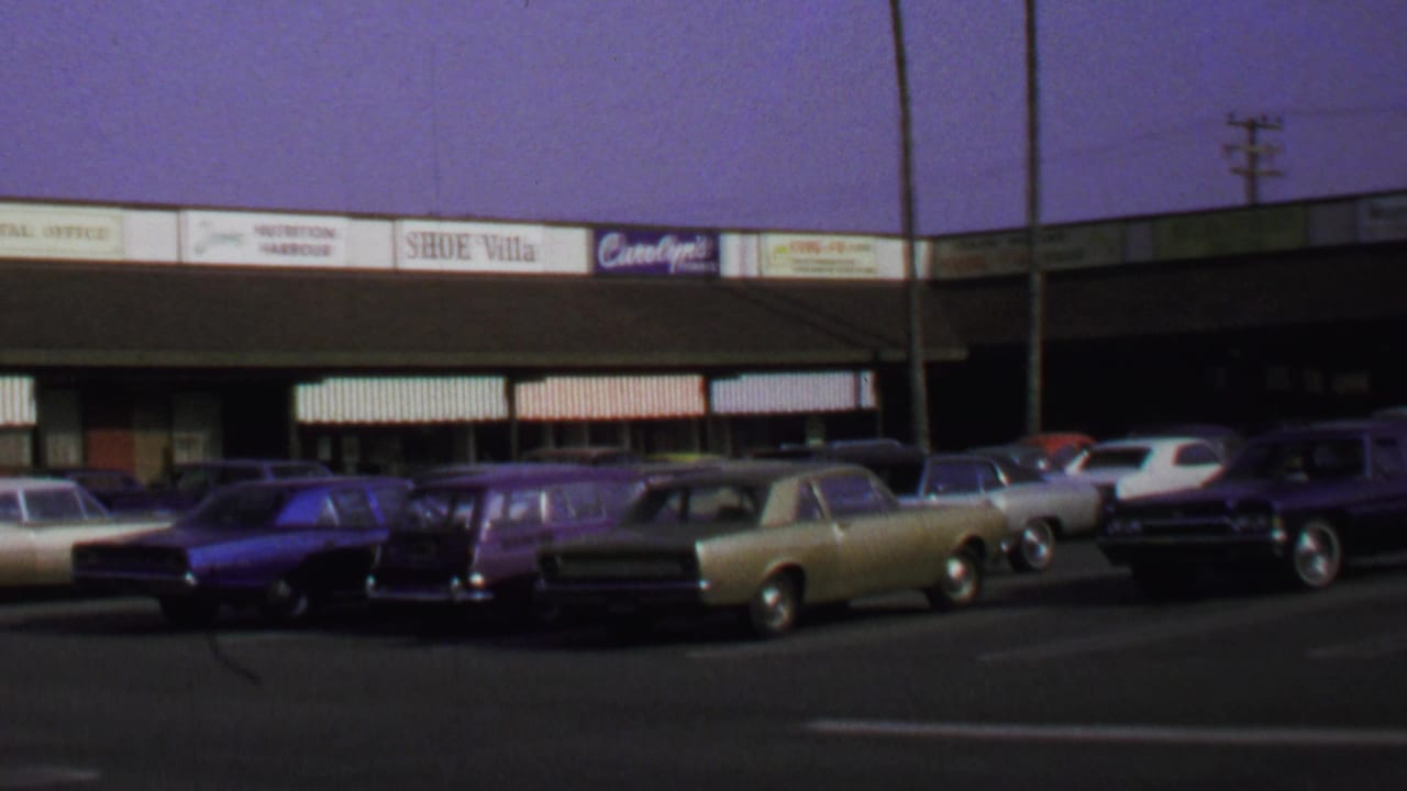 1969 Washington, DC home-movie view of parked late-1960s cars outside a low shopping center with visible storefront signs.