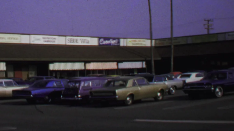 1969 Washington, DC home-movie view of parked late-1960s cars outside a low shopping center with visible storefront signs.