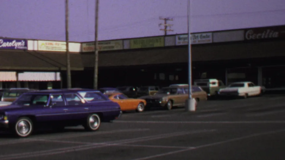1969 Washington, DC home-movie frame showing a purple wagon, orange car, and small shopping center storefronts.