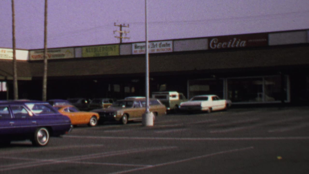 1969 Washington, DC home-movie frame showing a strip shopping center, parked classic cars, and visible storefront signage.
