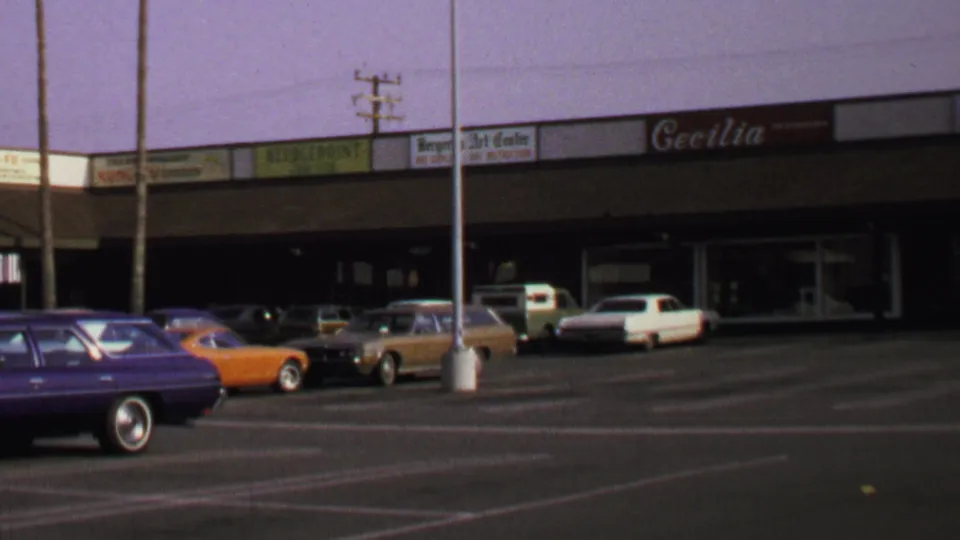 1969 Washington, DC home-movie frame showing a strip shopping center, parked classic cars, and visible storefront signage.