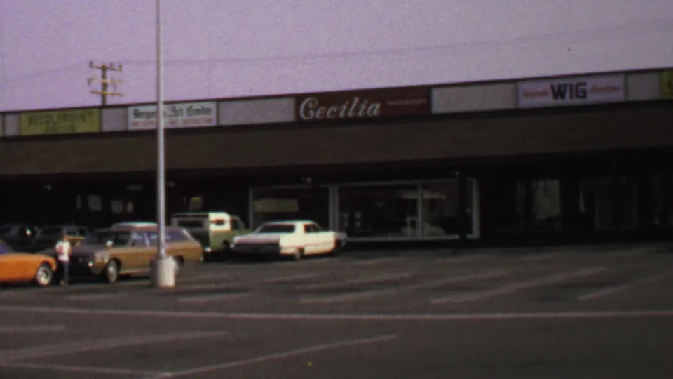 1969 Washington, DC frame showing a person crossing a shopping-center parking area near classic parked cars and storefront signs.