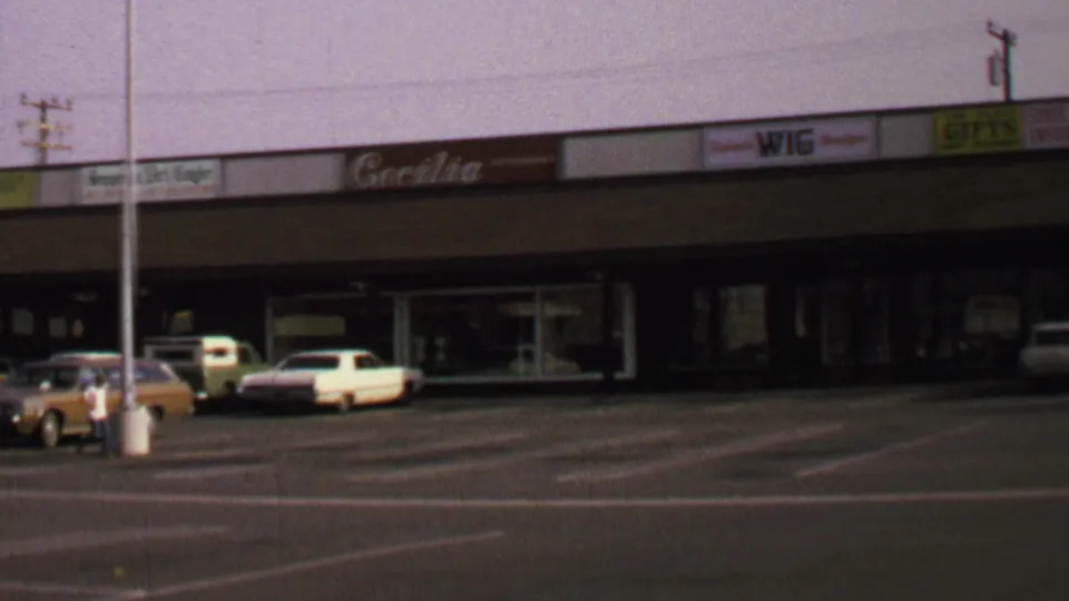 1969 Washington, DC home-movie still with storefront signs, parked classic cars, and an open shopping-center parking lot.