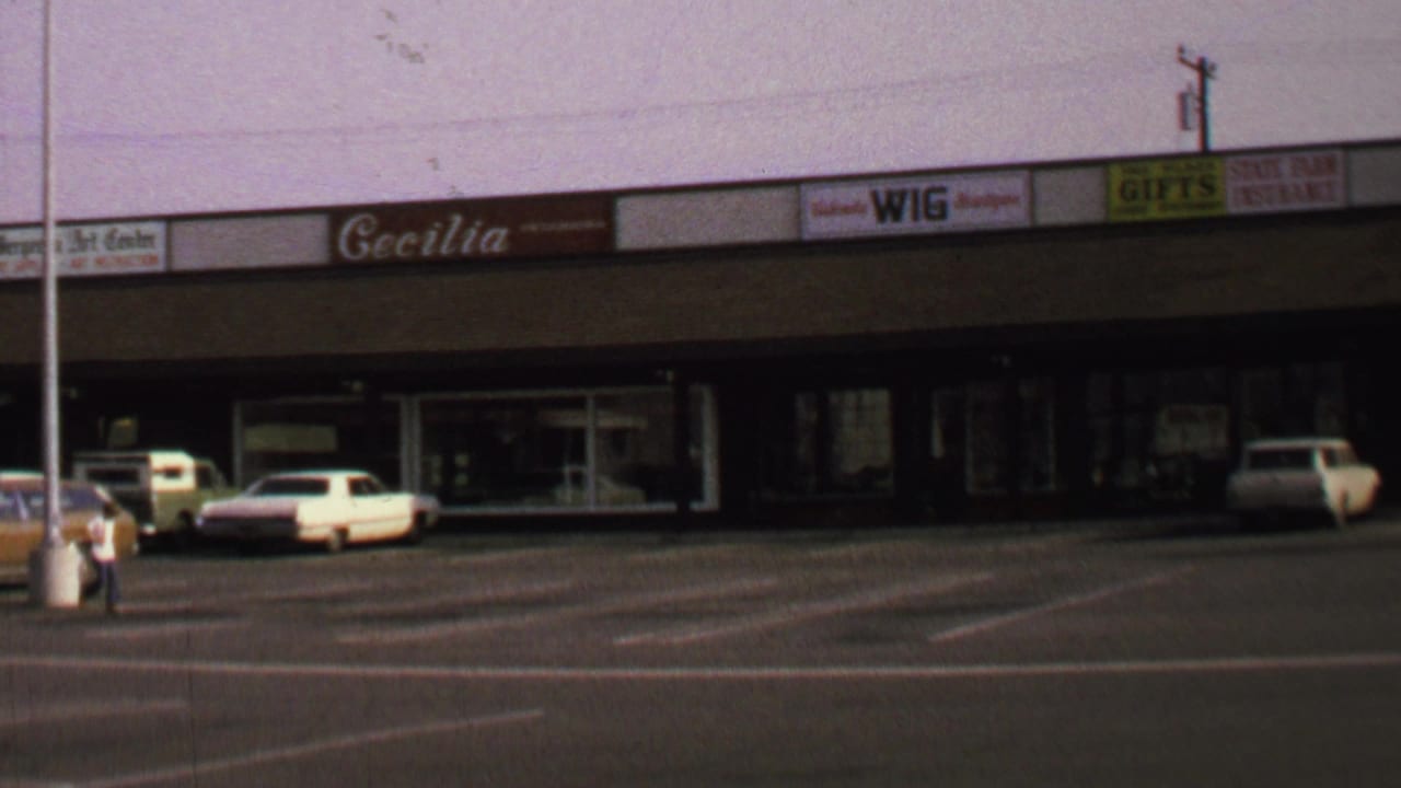 1969 Washington, DC home-movie frame showing a strip shopping center, wide car park, storefront signs, parked cars, and one distant pedestrian.