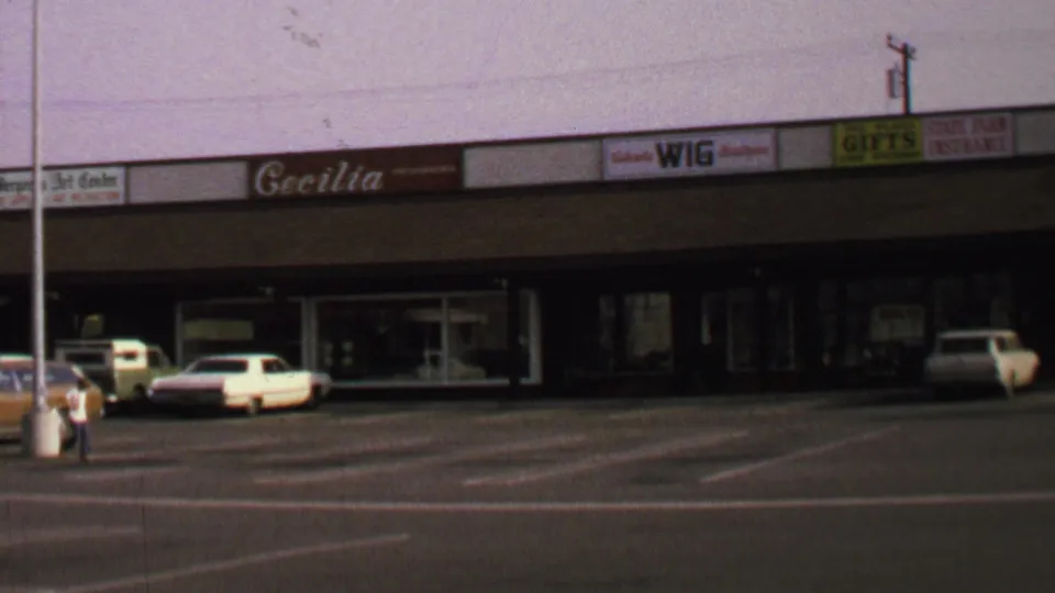 1969 Washington, DC home-movie frame showing a strip shopping center, wide car park, storefront signs, parked cars, and one distant pedestrian.