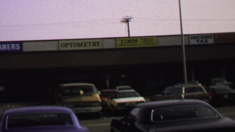 1969 Washington, DC frame focused on strip-mall signage above parked sedans and station wagons in a dim storefront row.