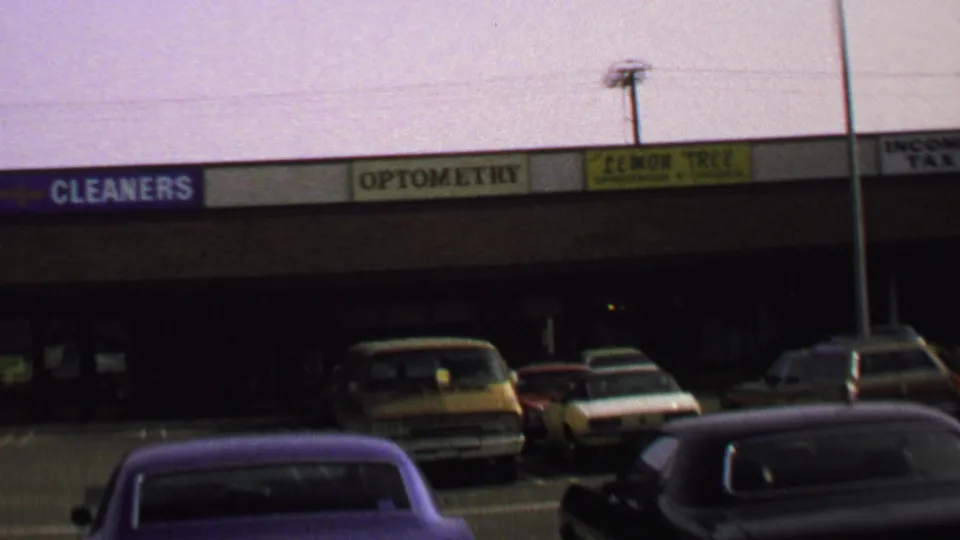 1969 Washington, DC home-movie view of a strip-mall facade with parked cars, signs, and a purple car entering the lower frame.