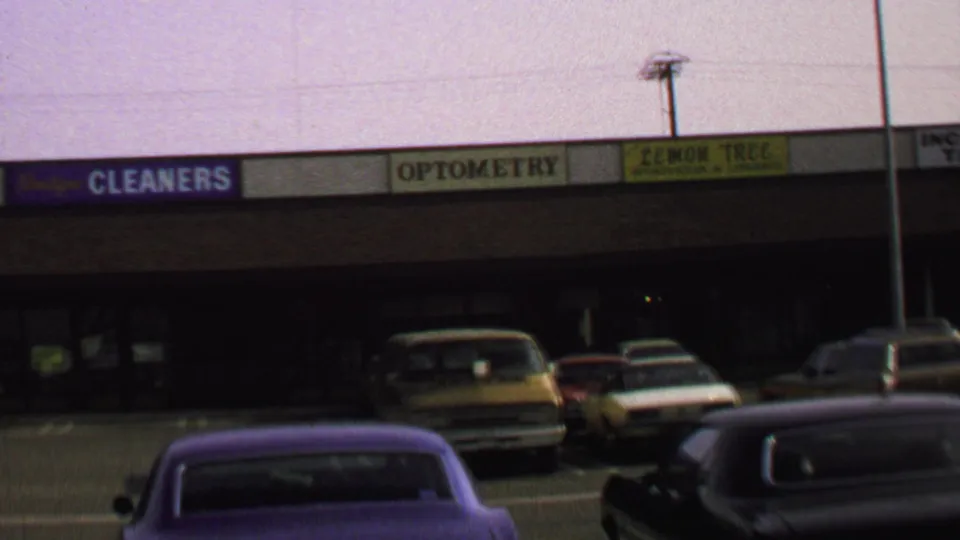 1969 Washington, DC frame showing a strip shopping center, visible service-business signs, parked cars, and a soft grainy 8mm look.