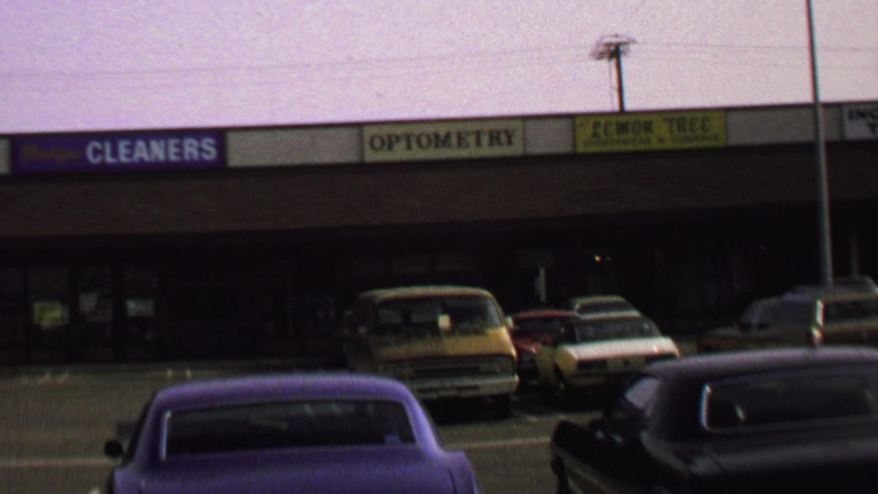 1969 Washington, DC home-movie frame showing a commercial plaza, parked period cars, and storefront signs for cleaners, optometry, and Lemon Tree.