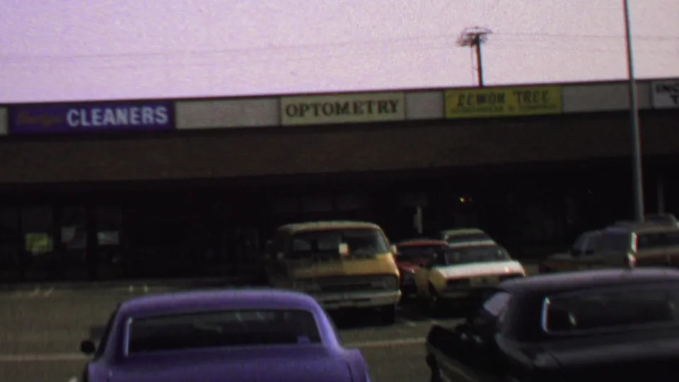 1969 Washington, DC home-movie frame showing a commercial plaza, parked period cars, and storefront signs for cleaners, optometry, and Lemon Tree.