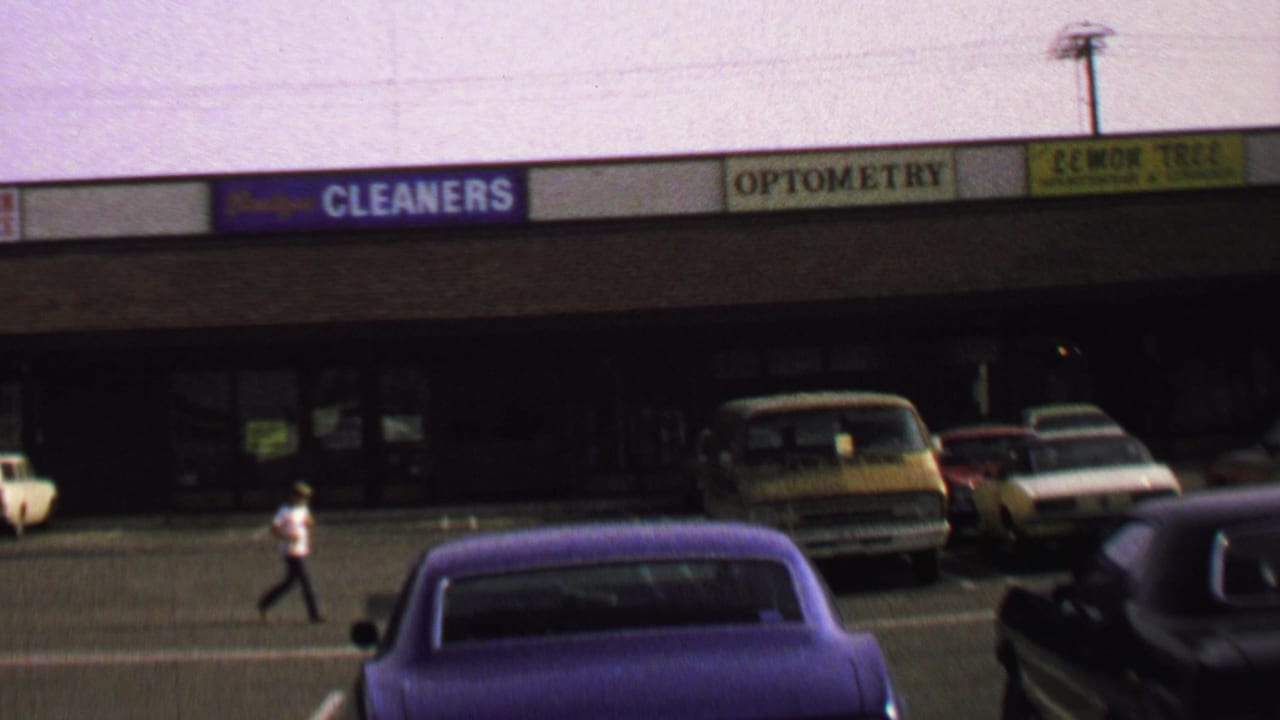 1969 Washington, DC frame with a pedestrian crossing a quiet commercial-plaza parking lot between parked cars and storefront signs.