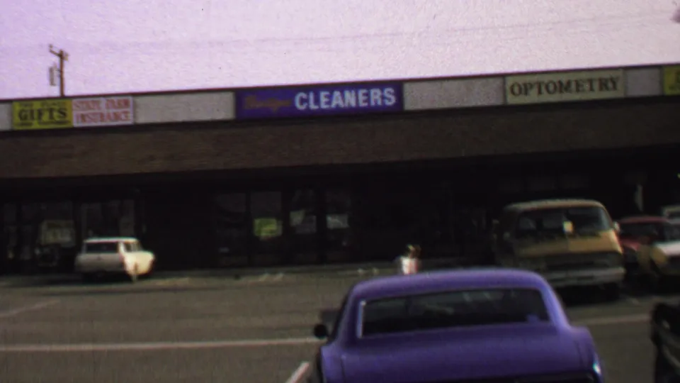 1969 Washington, DC home-movie view of a purple car in a commercial-plaza lot with cleaners, optometry, and small shop signs behind it.