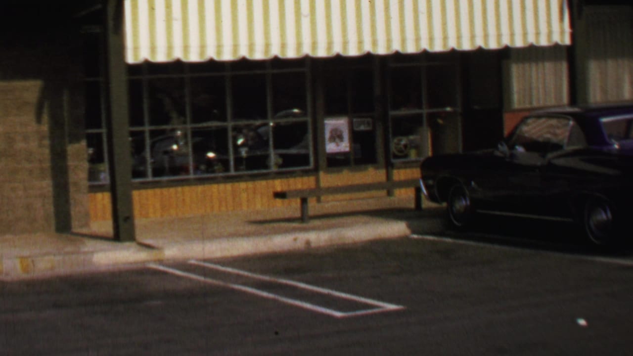 Dark classic car parked outside a glass storefront with striped awning and sidewalk bench.