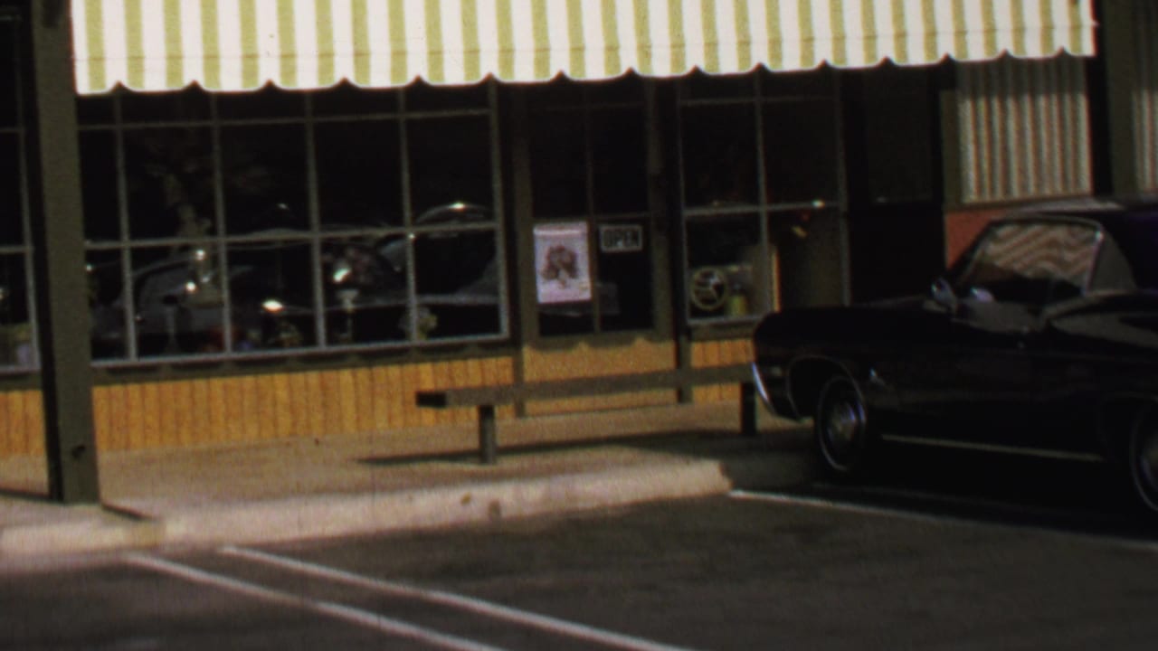 Washington, DC shop exterior from 1969 with striped awning, reflective window panes, sidewalk bench, and a parked dark car.