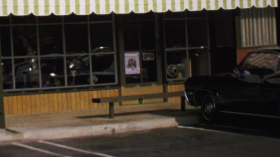 Closer 1969 Washington, DC view of the glass storefront, open sign, striped awning edge, bench, curb, and dark car front.