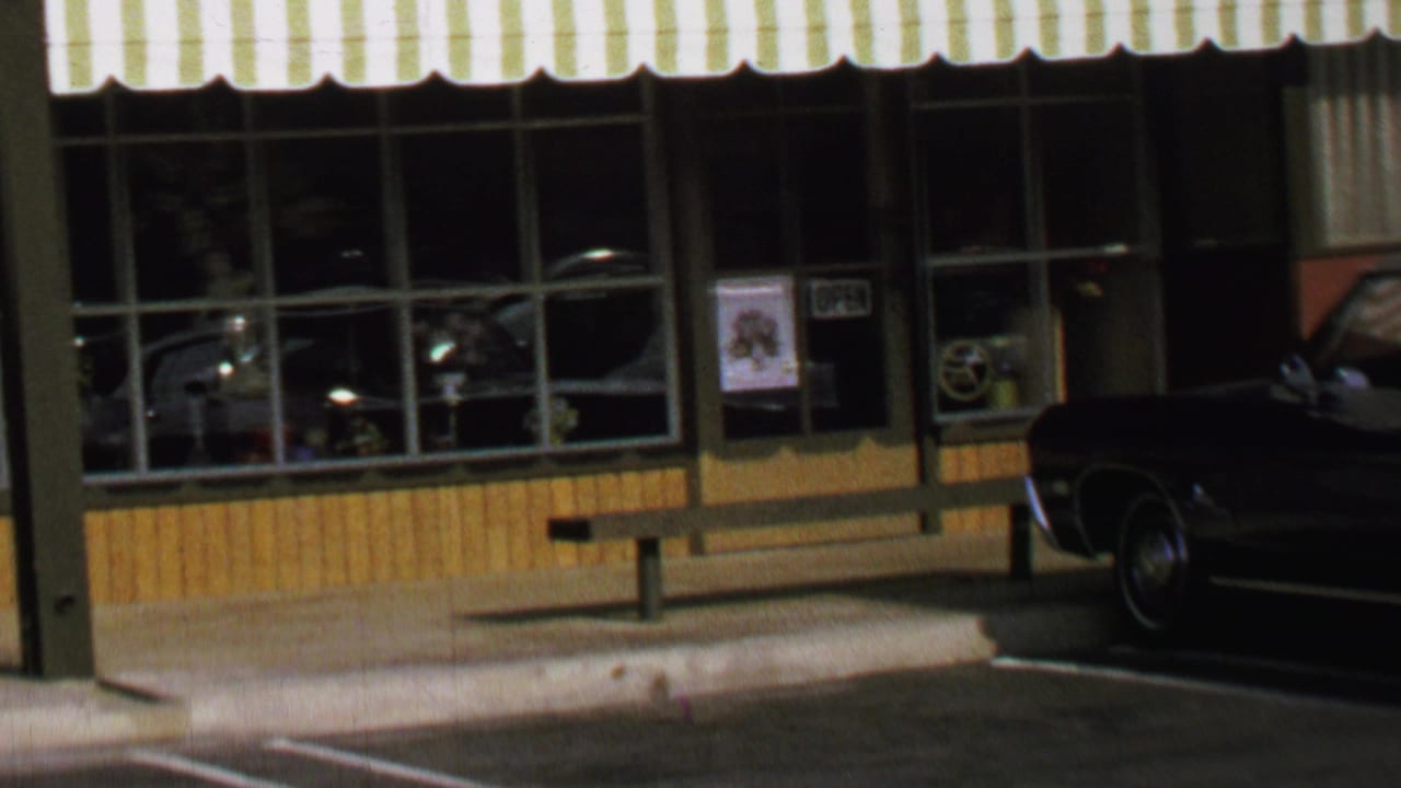 1969 Washington, DC storefront with striped awning, window grid, sidewalk bench, curb, parking lines, and a dark classic car partly visible.