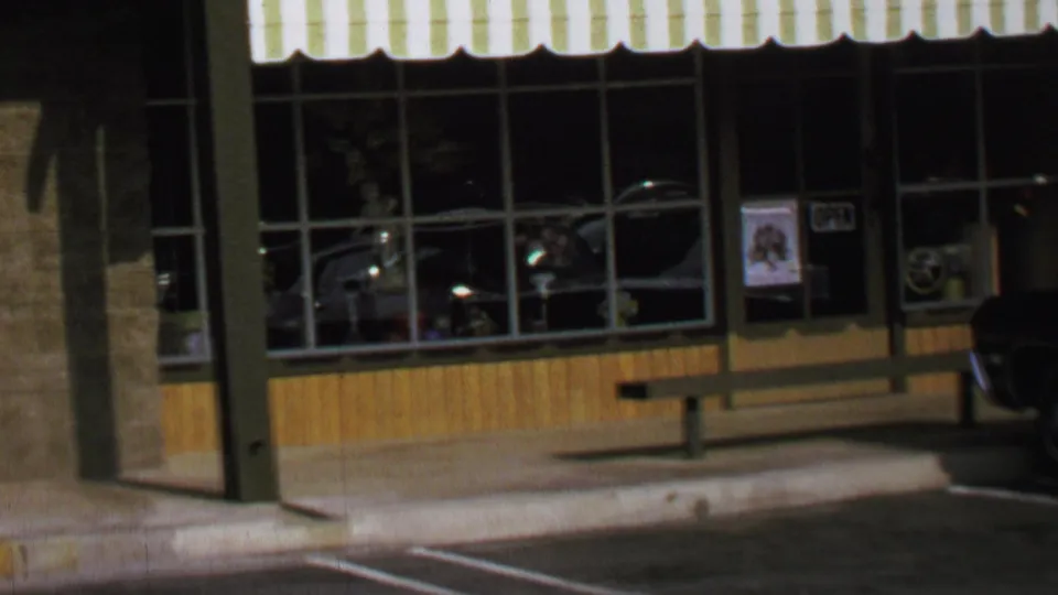 Static 1969 Washington, DC view of storefront windows, striped awning, sidewalk bench, curb markings, and a parked dark car at frame right.