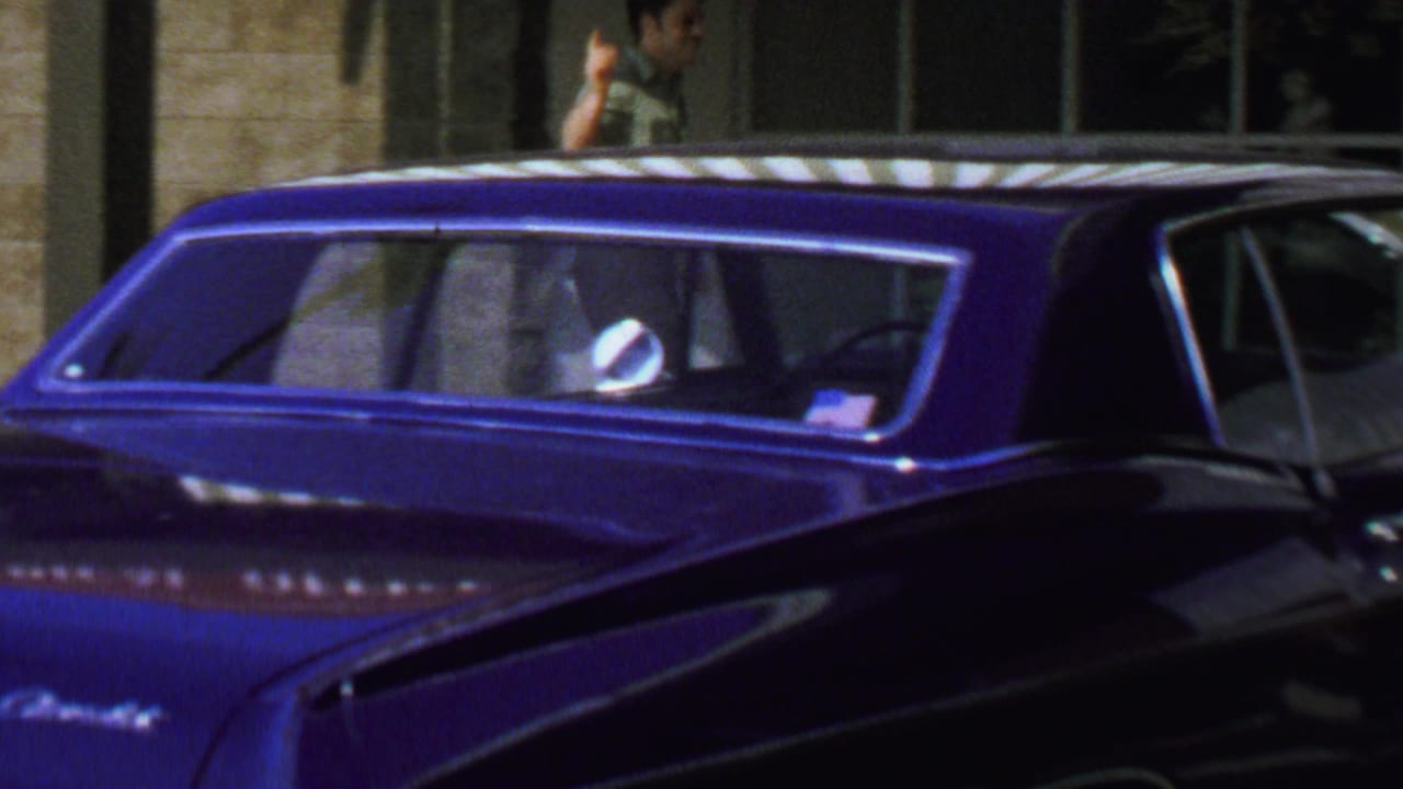 1969 Washington, DC home-movie frame showing a glossy blue classic car near storefront windows, with a young man partly visible behind the car.