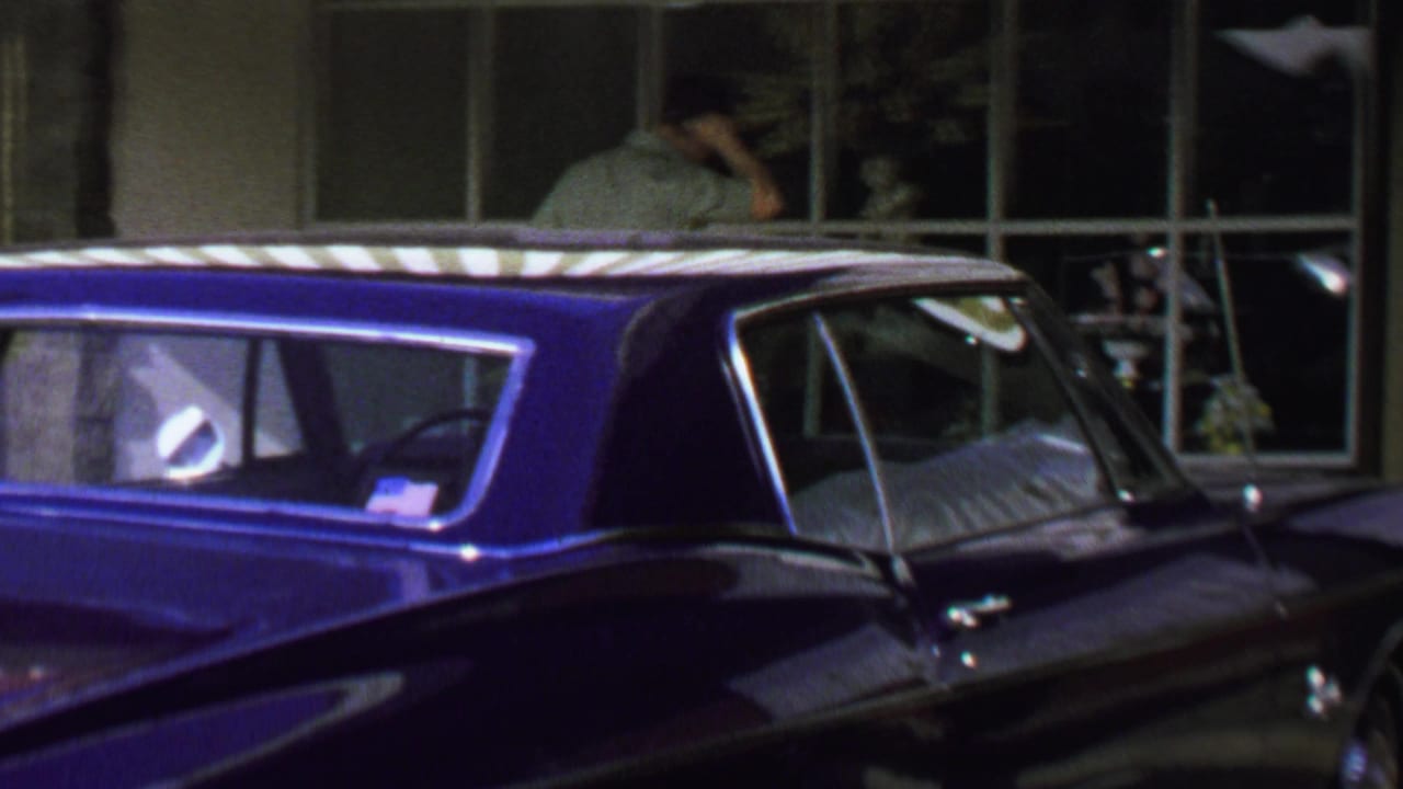1969 Washington, DC street frame with a blue classic car parked beside storefront windows and a man standing near the display area.