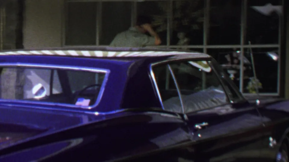 1969 Washington, DC street frame with a blue classic car parked beside storefront windows and a man standing near the display area.