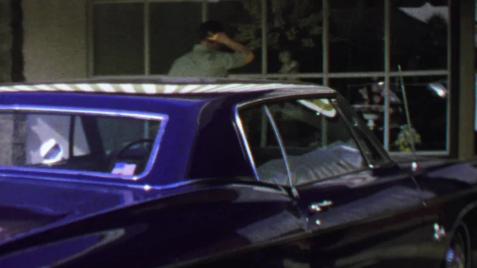 1969 Washington, DC home-movie frame showing a young man touching his head near a storefront, partly obscured by a blue parked car.