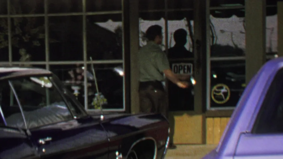 1969 Washington, DC home-movie frame showing the man at the shop door, with the OPEN sign and two parked cars partly visible.