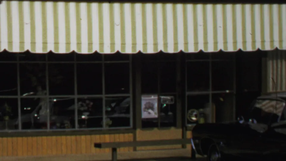 1969 Washington, DC storefront with striped awning, large windows, street reflections, and a dark car partly visible outside.