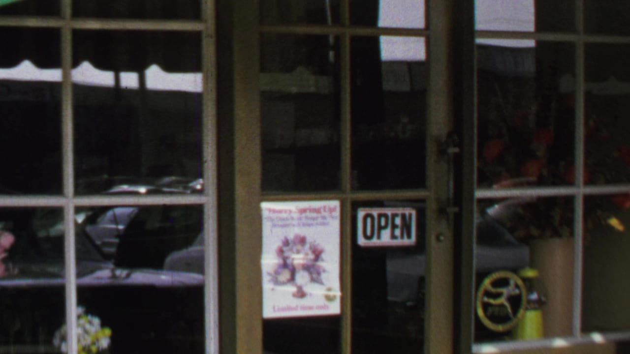 1969 Washington, DC storefront detail with a glass door, open sign, flower poster, reflected cars, and floral items inside.
