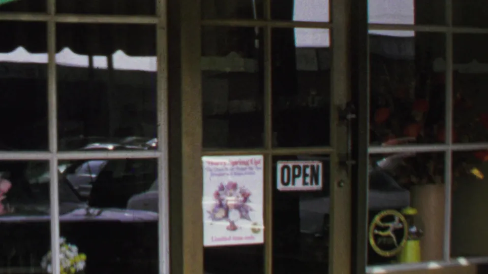 1969 Washington, DC storefront detail with a glass door, open sign, flower poster, reflected cars, and floral items inside.