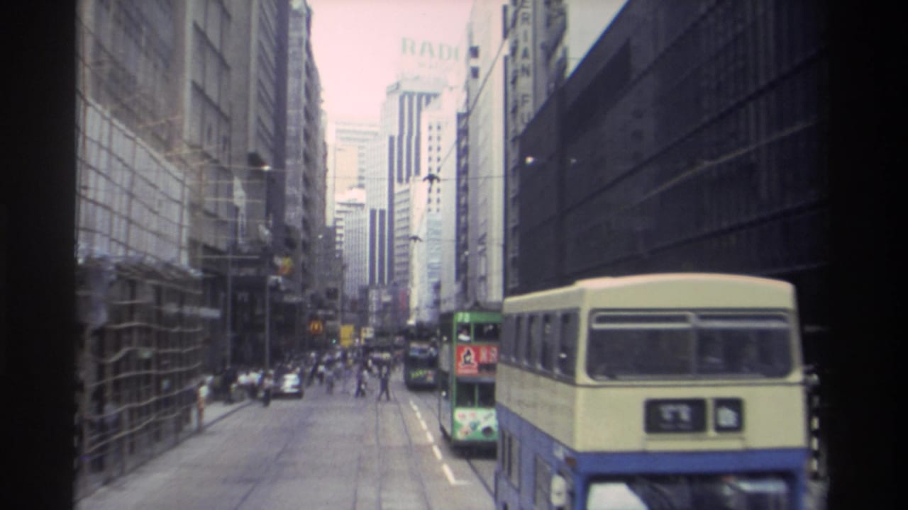 1982 Hong Kong street view with tall commercial buildings, double-decker buses, cars, and small pedestrian figures along the roadway.