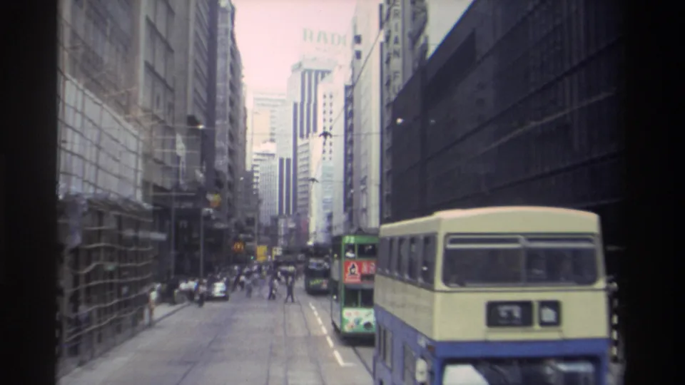 1982 Hong Kong street view with tall commercial buildings, double-decker buses, cars, and small pedestrian figures along the roadway.