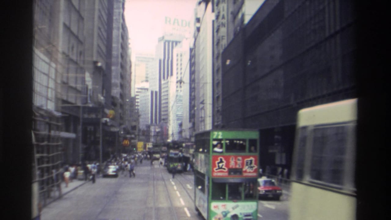 Hong Kong street traffic in 1982, with a green double-decker transit vehicle, a cream-blue bus, cars, pedestrians, and high-rise facades.