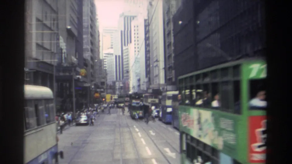 1982 Hong Kong city street with a nearby green transit vehicle, distant cars, pedestrians, road markings, and dense commercial buildings.