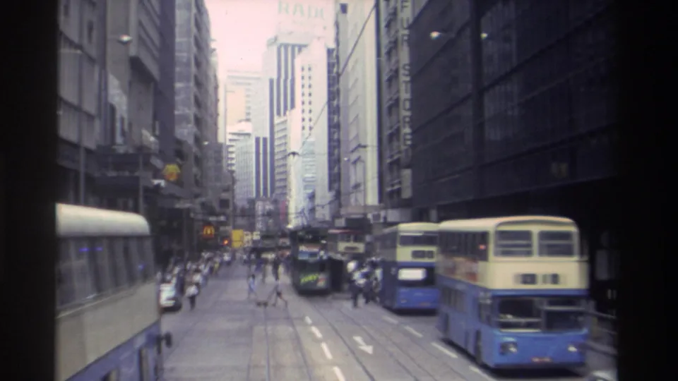 Hong Kong street scene from 1982 showing cream-blue double-decker buses, pedestrians crossing, cars, tall buildings, and commercial signage.
