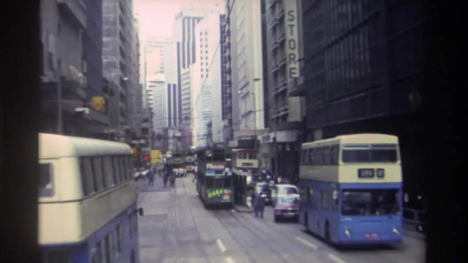 Moving 8mm view of a Hong Kong street in 1982, with double-decker buses, cars, pedestrians, overhead wires, and vertical signs.