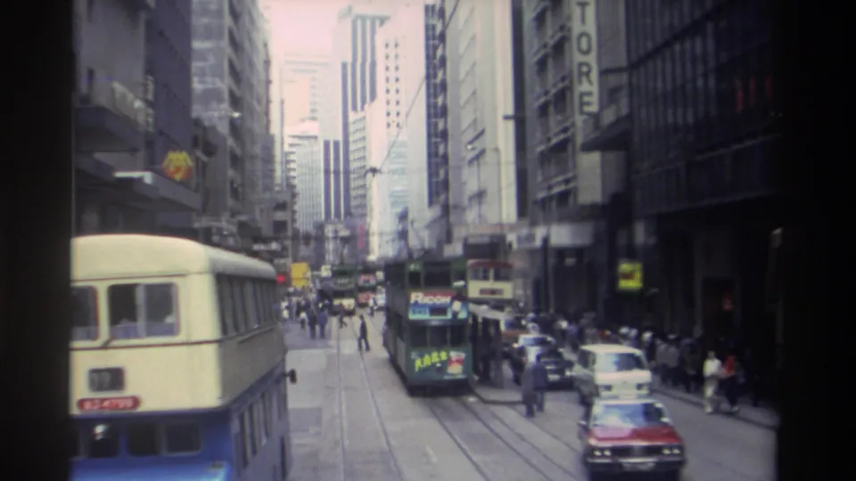 1982 Hong Kong traffic scene showing a bus in the foreground, a green tram ahead, cars at the curb, and pedestrians along commercial buildings.