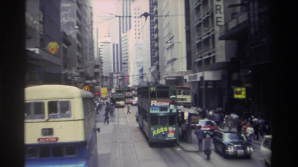 Hong Kong in 1982 seen from a moving vehicle, with a green tram centered among buses, cars, pedestrians, tall buildings, and shop signage.