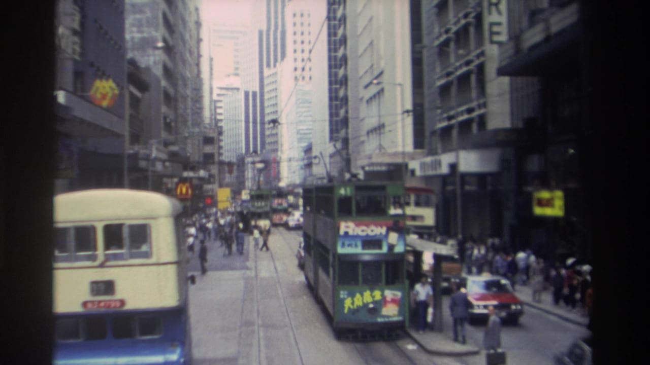 1982 Hong Kong street view with a cream and blue double-decker bus, a green tram, pedestrians, traffic, and commercial signs.