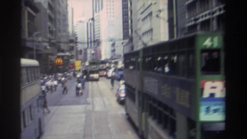 1982 Hong Kong street scene with a green double-decker tram passing near the camera, traffic ahead, pedestrians, and tall commercial buildings.