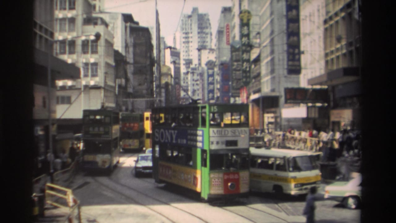 Green double-decker tram and cars on a narrow Hong Kong street with tall buildings and signs.