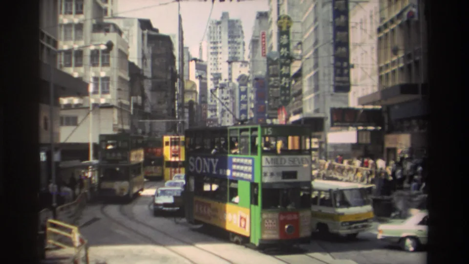 Green and yellow double-decker tram in 1982 Hong Kong traffic, with cars, pedestrians and vertical Chinese signage visible.